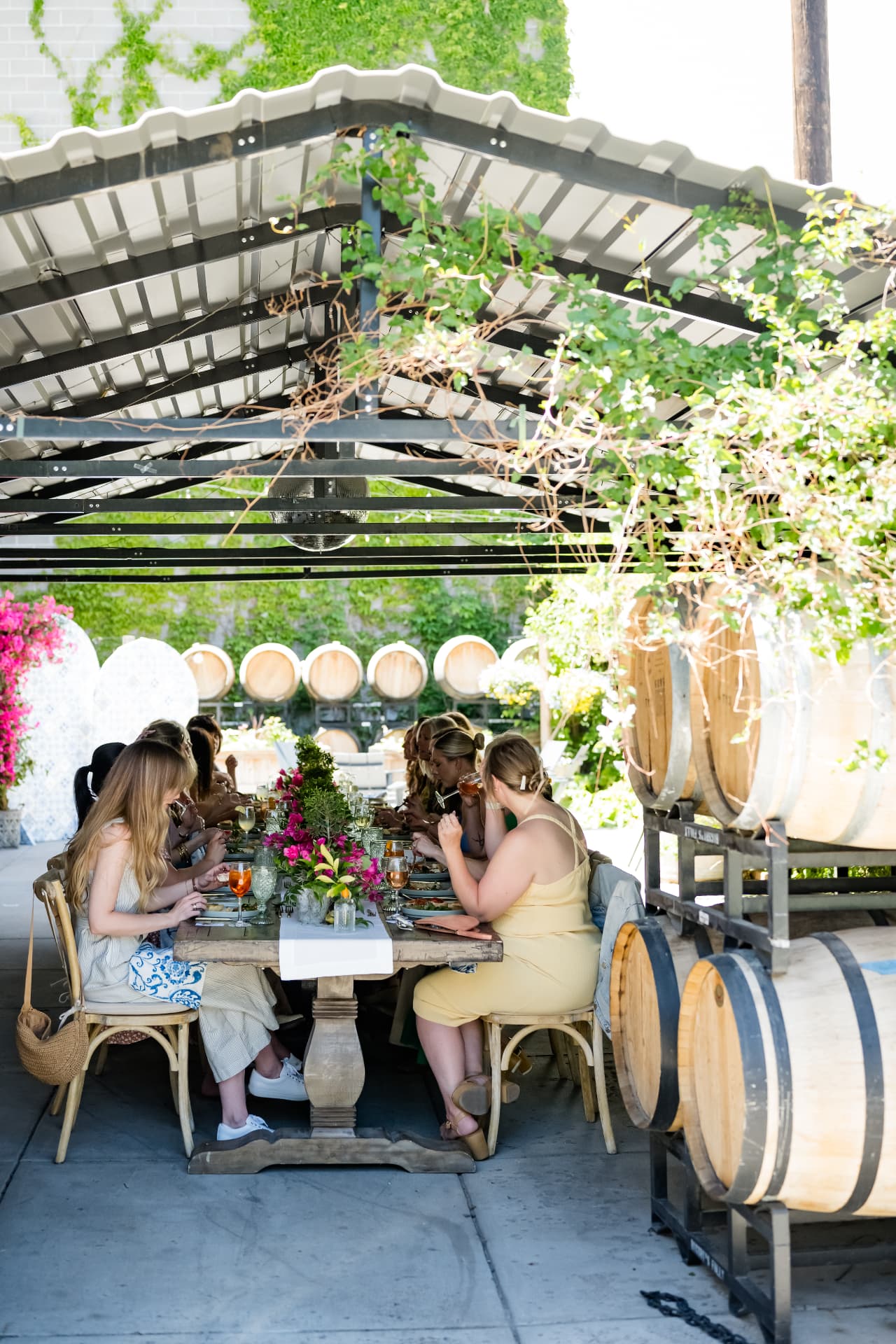 Guests dining at a flower-filled table beneath the ivy-covered courtyard canopy beside wine barrels